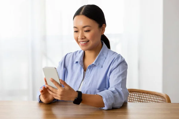 Smiling attractive millennial asian woman wearing casual outfit sitting at desk at home, using phone, reading news or email, texting friends, scrolling while resting at weekend, copy space