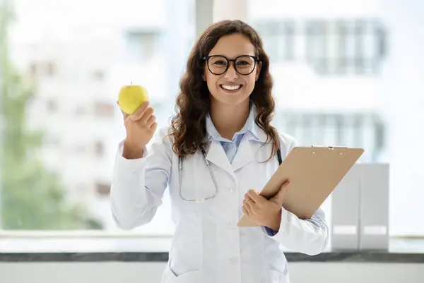 Friendly attractive young woman wearing white medical coat and eyeglasses doctor general physician standing next to window at clinic, holding clipboard, showing apple, recommending healthy diet