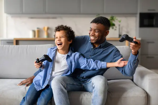 Exuberant black preteen boy and his father sharing joyful moment, laughing and playing video games together, creating memories in their cozy living room