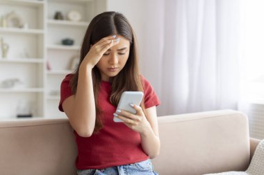 Endişeli genç Asyalı kadın cep telefonunda mesaj okuyor, Koreli kadın endişeli ya da kafası karışık, evdeki oturma odasında kanepede oturuyor, boşluğu kopyalıyor.