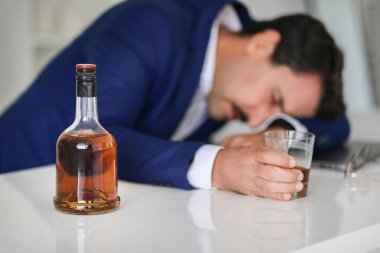 Mavi takım elbiseli bitkin ve sıkıntılı beyaz bir işadamı kafasını bir masanın üzerinde dinlendiriyor. Neredeyse dolu bir içki şişesi ve boş bir bardağın yanında. Muhtemelen stres veya tükenmişlik belirtisi.