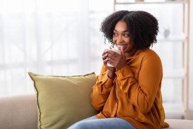 Mutlu bir şekilde gülümseyen, doğal kıvırcık saçlı Afro-Amerikan bir kadın, yeşil bir yastığın yanında uzanırken canlı bir lacivert bluz giymiş beyaz bir kupa tutuyor.