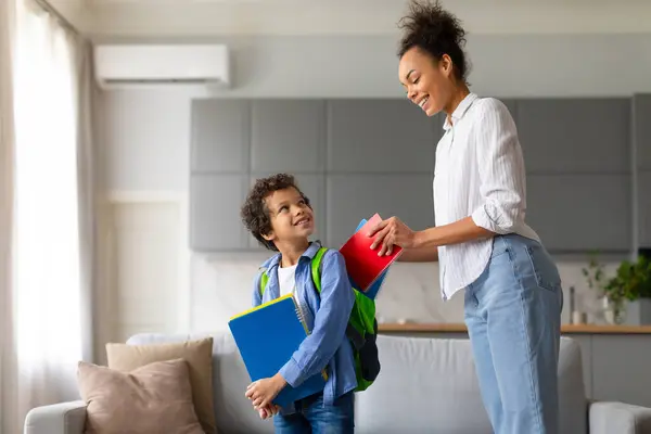 快乐的黑人妈妈把红皮笔记本放在她快乐的卷发儿子身上 她背着背包 准备在明亮的客厅里迎接新的一天 — 图库照片