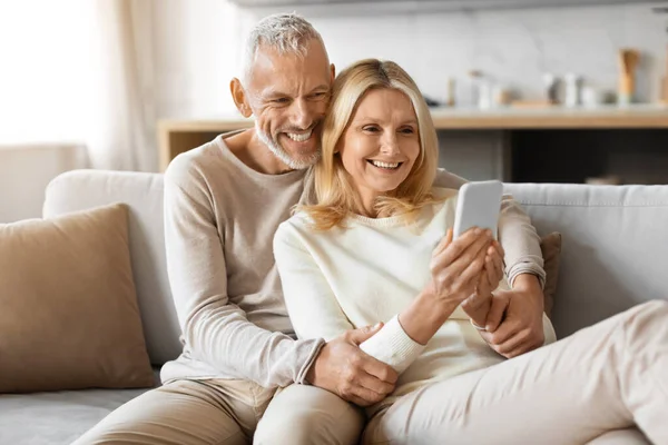Happily married senior couple relaxing on couch at home, embracing, scrolling on social media, checking their kids or friends photos, using phone, websurfing, shopping online, copy space