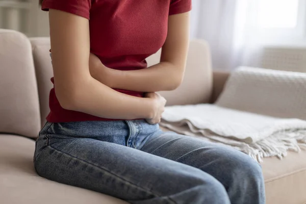 Uncomfortable young woman embracing her abdomen, feeling acute stomach ache while sitting on couch in well-lit living room, unrecognizable female suffering digestion problems, cropped shot