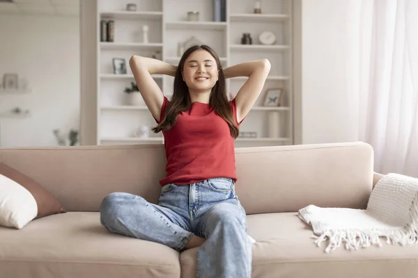 Smiling young asian woman enjoying moment of relaxation at home, beautiful korean lady sitting with eyes closed, stretching her arms behind her head and leaning back on soft beige sofa in living room