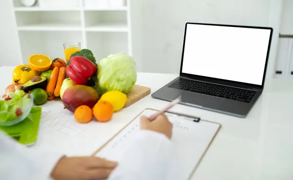 不知名的女医生会与病人进行在线咨询 坐在办公桌前 吃新鲜的有机水果和蔬菜 用空白屏幕看笔记本电脑 做笔记 — 图库照片