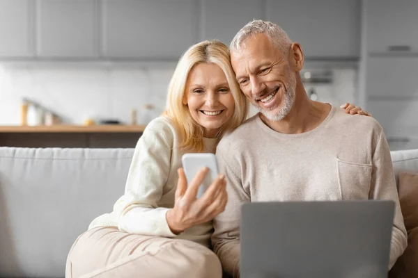 Cheerful retired couple using gadgets while relaxing together at home cozy studio apartment, sitting on couch, embracing, using smartphone and computer laptop, sharing nice content, copy space