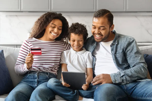 Cheerful Black Family Shopping Online With Digital Tablet And Credit Card At Home, Happy African American Parents And Their Little Son Relaxing On Couch With Modern Gadget, Purchasing In Internet