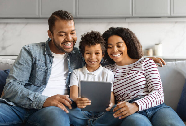 Black family with preteen son smiling joyfully while using digital tablet together, happy african american parents and male kid sitting on comfortable couch at home, relaxing with modern gadget