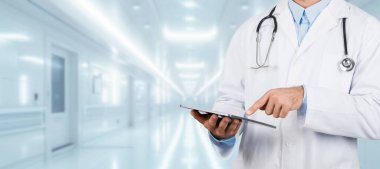 Boynunda steteskop olan beyaz önlüklü profesyonel Avrupalı bir doktor, aydınlık bir hastane koridorunda, panorama, kesilmiş bir tablet kullanıyor.
