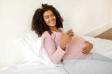 Güzel siyahi hamile bayan evde akıllı telefonuyla vakit geçiriyor, internete bakıyor ya da arkadaşlarıyla mesajlaşıyor, Afro-Amerikan hamile anne rahat bir yatakta dinleniyor, boşluğu kopyalıyor.
