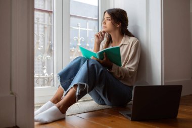 Düşünceli genç bir kadın elinde defterle pencereden dışarı bakıyor, düşünceli bir kadın yanında dizüstü bilgisayarla yerde oturuyor, huzurlu ışık dolu bir odada, üretkenlik telkin ediyor, boş alan yaratıyor.