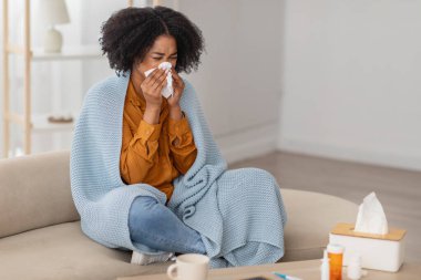 Kıvırcık saçlı Afrikalı genç bir Amerikalı kadın, evdeki bir kanepede oturmuş, yanında soğuk algınlığı ilacı ve kupası ile sıcak mavi bir battaniyeye sarılmış bir şekilde mendile hapşırıyor.