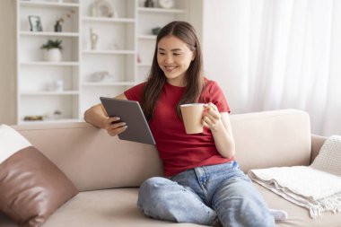 Dinlenmiş genç Asyalı kadın elinde dijital tablet ve kahve kupası, mutlu bin yıllık Koreli kadın oturma odasında kanepede dinleniyor, internete bakıyor ve sıcak içeceğin tadını çıkarıyor, uzay fotokopisini çekiyor.
