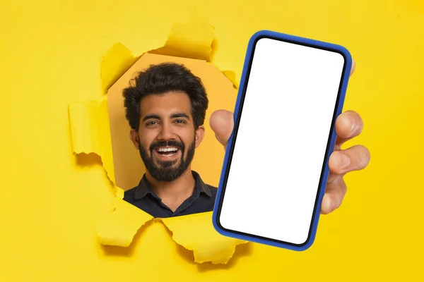 快乐的阿拉伯人展示手机 手臂伸向被撕破的床单 在黄色的工作室背景上展示带有空屏幕的大型智能手机 模仿手机的报价广告 — 图库照片