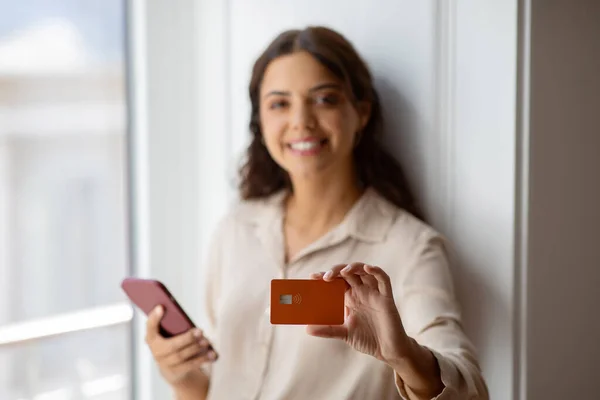 Confident young woman presenting credit card while holding smartphone, smiling beautiful lady standing by window at home, suggesting seamless online transaction, selective focus on hand with card