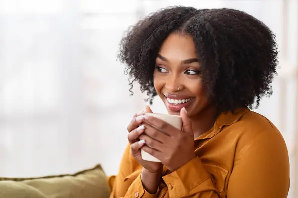Close-up of a radiant African American woman with a captivating smile, holding a mug with both hands, dressed in a mustard blouse, creating a feeling of warmth and comfort
