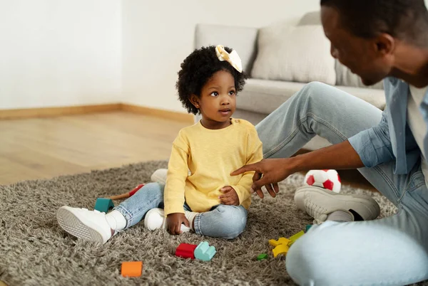 Loving african american dad teaching his toddler daughter to talk. Black man father sitting on carpet floor at home, communicating with his child little girl, playing with toys, copy space