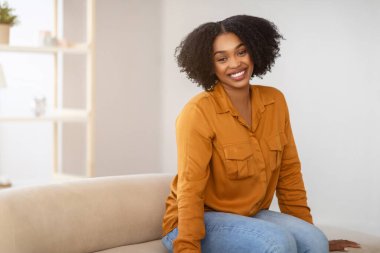 Neşeli, güzel, Afro-Amerikan bir kadın. Işıl ışıl gülümseyen, hardal sarısı bir tişört giyen, bej renkli bir kanepeye oturmuş, ev içinde rahat bir odada dinleniyor.