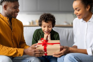 Sevecen siyah çocuk, sevgi dolu ailesinden hediye alırken gözleri şaşkın bir şekilde evde iç açıcı bir aile sahnesi yakalıyor.