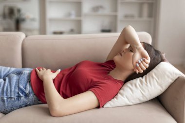 Genç Asyalı bir kadın, bir eli karnında, diğeri alnında kanepede yatarken midesinde rahatsızlık hissediyor. Hasta Koreli bir kadın karın ağrısı çekiyor, acı içinde.