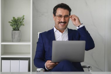 Mavi takım elbiseli, kendinden emin ve neşeli Kafkasyalı olgun iş adamı modern bir ofiste dizüstü bilgisayar üzerinde çalışırken iç dekorasyonu minimalist olan gözlük ayarlıyor. Cihazla iş, iş