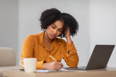 Konsantre olmuş, kıvırcık saçlı, hardal rengi gömlek giyen, laptopun yanına notlar yazan ve ofisteki ahşap masaya kahve fincanı koyan mutsuz, genç, Afro-Amerikan bir kadın.