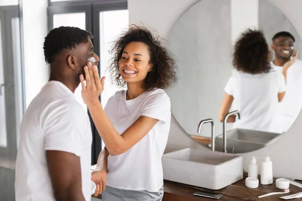 笑顔のアフリカ系アメリカ人のカップルは 自宅のバスルームに立って 楽しいスキンケアのルーチンを共有しています 妻は保湿剤を保持し 夫に優しく顔を合わせ 愛情と親密な瞬間を作成する — ストック写真