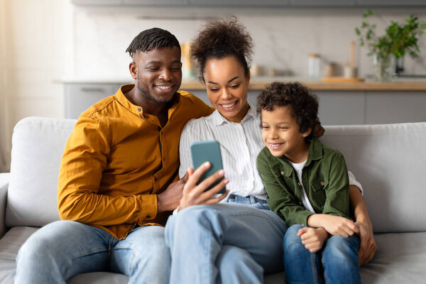 Warm and engaging black family of three shares fun moment as they look at smartphone together, smiling and enjoying each others company, sitting on sofa