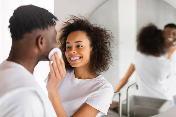 Happy African American couple engages in pampering and facial skincare in their bathroom, wife applying moisturizer or lotion to husbands face using cotton pad, moisturizing skin after shaving