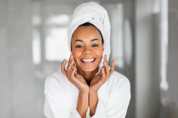 Happy attractive African American lady with glowing complexion carefully applies moisturizer to face, enjoying facial skincare routine, personal pampering and wellbeing in modern bathroom