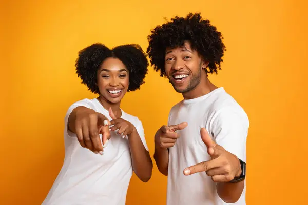Happy cool millennial african american couple pointing and smiling at camera. Cheerful young black man and woman in white t-shirts gesturing isolated on colorful studio background
