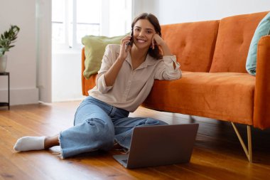 Mobil İletişim. Cep telefonuyla konuşan ve evinde dizüstü bilgisayar kullanan gülümseyen genç kadın, mutlu bir kadın telefon konuşmasının keyfini çıkarıyor, oturma odasında yerde oturuyor.