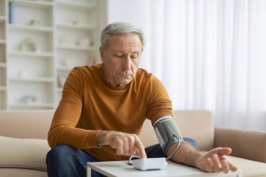 Kanepede oturan yaşlı bir adam modern elektronik tonometreyle kan basıncını ölçüyor. Dedem uyandıktan sonra sabah check-up yapıyor ya da kendini hasta hissediyor.