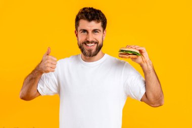 Abur cuburu severim. Cheeseburger tutan neşeli sakallı adamın portresi ve sarı stüdyonun arka planında kameraya gülümseyen baş parmaklar. Favori fast food 'un tadını beğenen bir yiyici