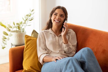 Evdeki koltukta dinlenirken cep telefonuyla konuşan güzel genç bir bayan, mutlu gülümseyen bir milenyum kadını, keyifli cep telefonu sohbetleri, oturma odasındaki koltukta oturan...