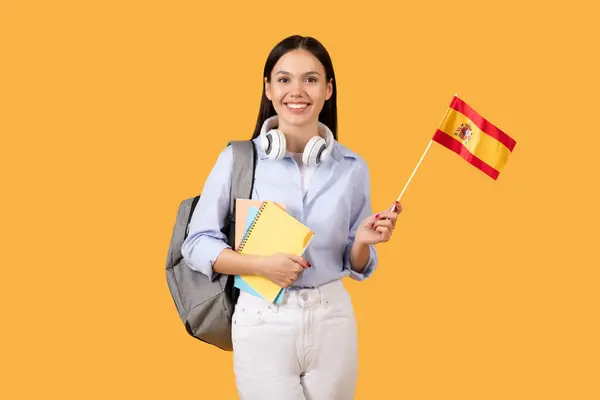 小さなスペインの旗を持っているヘッドフォンとバックパックを持つ笑顔の女性学生 手元にノートブック 黄色の背景にポーズ — ストック写真