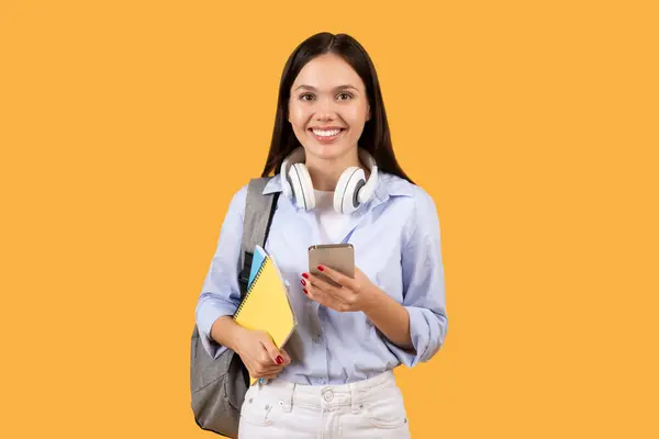 穿着蓝色衬衫 脖子上戴着耳机 手持智能手机和黄色笔记本 背对着黄色背景的年轻女性 — 图库照片