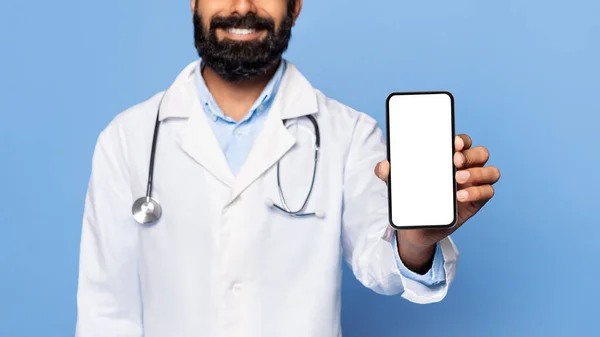 笑印度男医生风趣的胡子 展示智能手机与空白屏幕 身穿白色实验室外套和听诊器 蓝色背景 医疗广告 — 图库照片