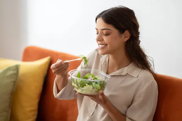 Relaxed young woman eating fresh green salad from bowl at home, happy millennial lady comfortably sitting on orange sofa in sunny living room, embodying healthy lifestyle, free space