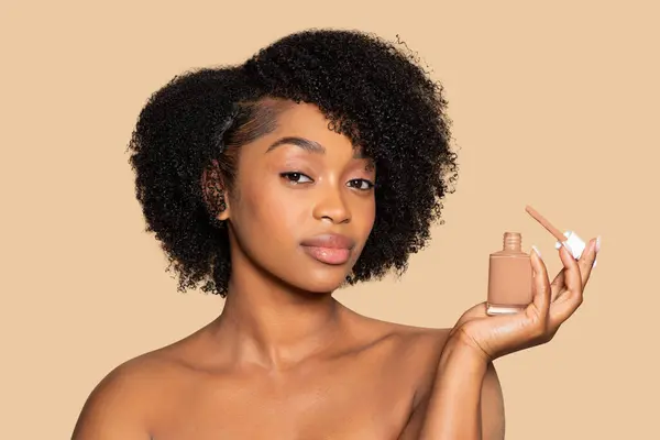 stock image Serene African American woman with curly hair displaying bottle of liquid foundation, highlighting the perfect match for her skin tone against smooth beige backdrop