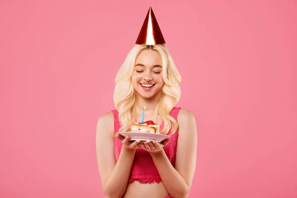 パーティー帽子のヨーヨーヨーの若いブロンドの女性 目を閉じて大切な瞬間 穏やかにライトキャンドルで誕生日ケーキを保持 ピンクの背景に対して — ストック写真