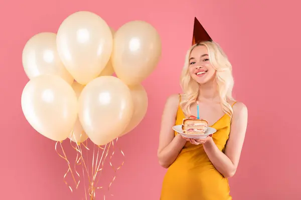 パーティー帽子と黄色のドレスで陽気なブロンドの女性 ライトキャンドルと誕生日ケーキを開催 クリーム風船の束を伴う ピンクの背景 — ストック写真