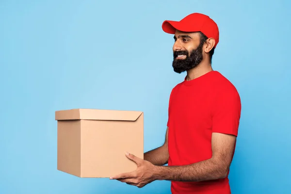 Happy delivery indian man with beard in red uniform and cap carrying cardboard box, holding package, standing against solid blue background