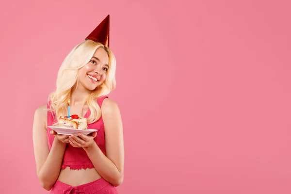 パーティー帽子のハッピー若いブロンドの女性 キャンドルで誕生日ケーキのスライスを保持 喜んで笑顔で自由なスペースで一見 ソフトピンクの背景に対して — ストック写真