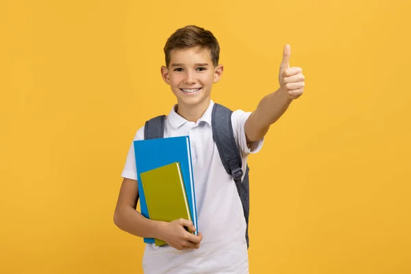 カメラで親指を示すバックパックと本 明るい黄色のスタジオの背景に立って 教育について楽観主義を表現する幸せなティーンエイジャーの男の子 コピースペース — ストック写真