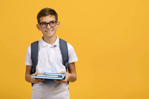 ガラスとバックパックを身に着けているインテリジェントなスクールボーイは 教科書を保持し コピースペースを見下ろし 学校の準備ができている笑顔の男性の子供 イエロースタジオの背景に立って パノラマ — ストック写真
