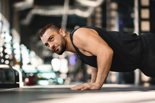 Bearded man holding plank position while training at gym, handsome young male athlete demonstrating core strength and focus during his fitness routine in modern sport club, side view