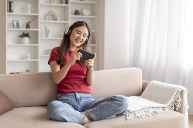 Akıllı telefonuyla video oyunu oynayan neşeli, genç Koreli kadın koltukta dinlenirken, kulaklık takan mutlu Asyalı kadın oturma odasında kanepede dinleniyor, modern teknolojilerin tadını çıkarıyor.